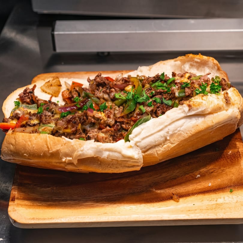 A close-up of a loaded cheesesteak sandwich with peppers, onions, and melted cheese on a wooden board.
