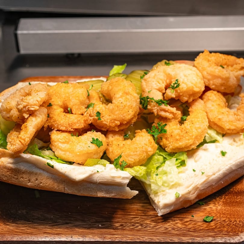 Close-up of a shrimp po'boy sandwich with fried shrimp, lettuce, pickles, and sauce on a wooden board.