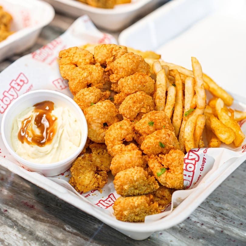 A Holy Fry takeout container filled with crispy fried shrimp, seasoned fries, and a side of dipping sauce.